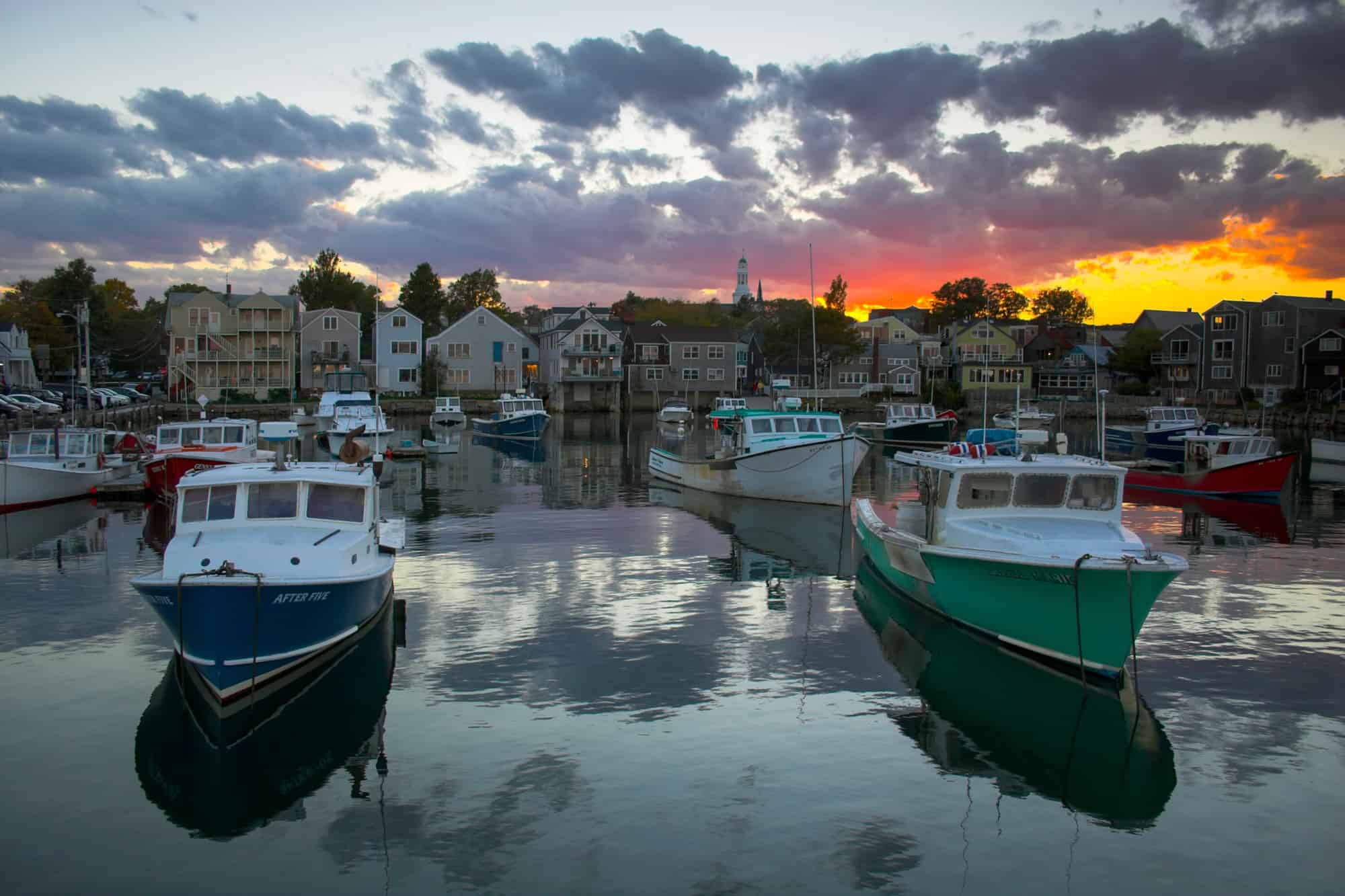 boats sit in the rockport harbor at sunset as orange light spreads along the sky among dark clouds