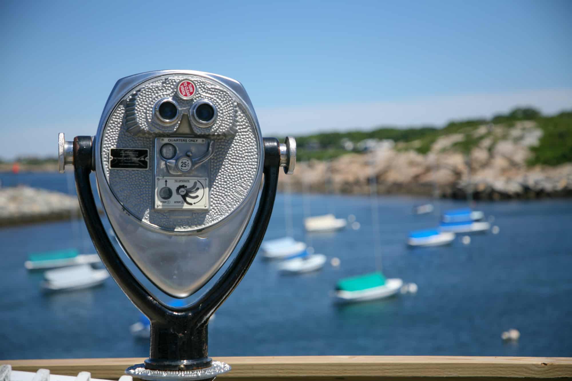 a viewfinder sits along the coast to take a closer look for a quarter, behind you can see boats floating in the water along the rockport, massachusetts, coastline