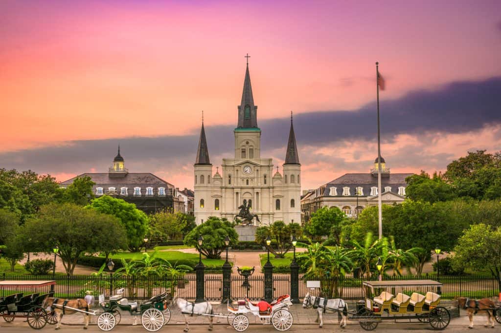 jackson square in new orleans with a row of horse-drawn carriages out front of a large catedral under a pink and orange sky