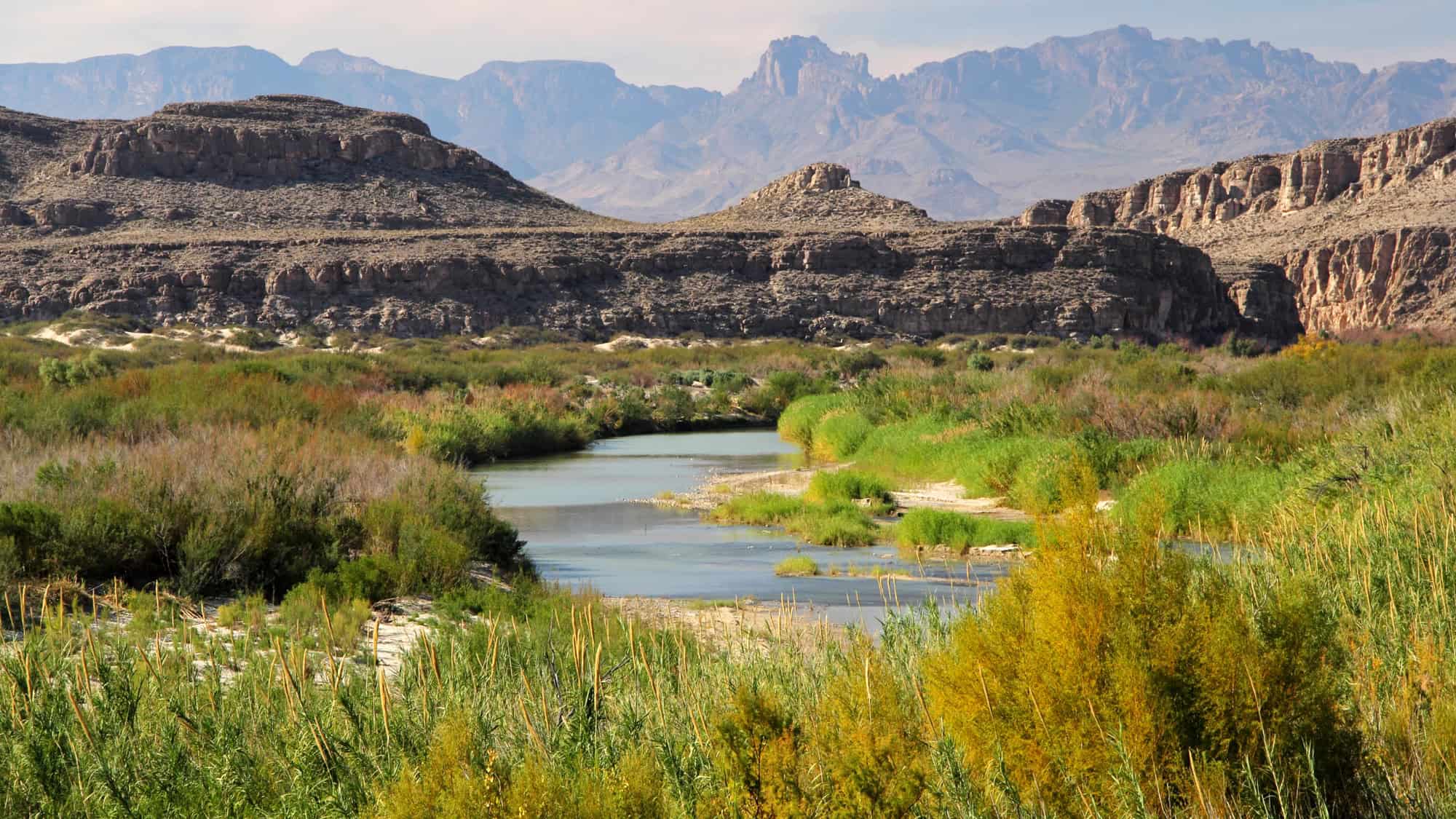 A winding river flows through green vegetation and rocky cliffs in a desert mountain landscape.