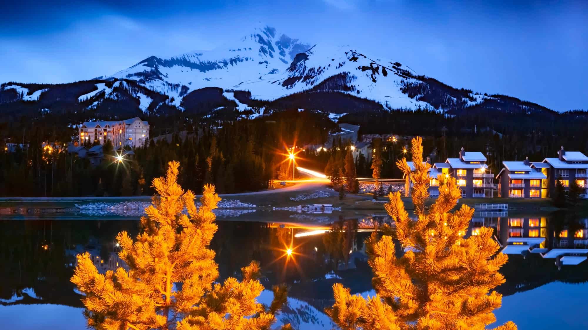 Snow-covered peaks rise behind a warmly lit mountain resort, with lights reflecting in a calm lake.