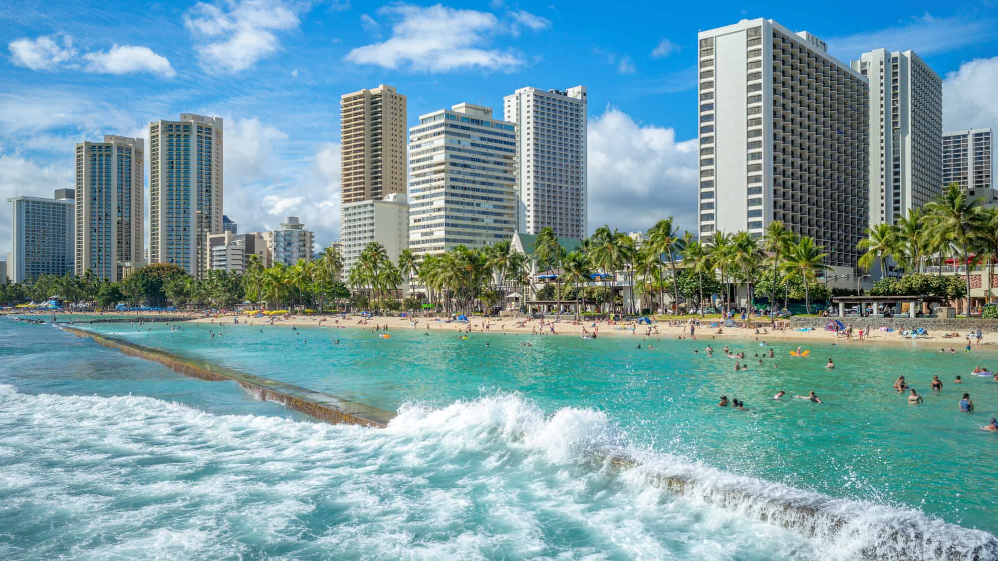 Turquoise waves crash against a barrier as people swim and relax on the sandy beach in front of high-rise hotels in Honolulu.