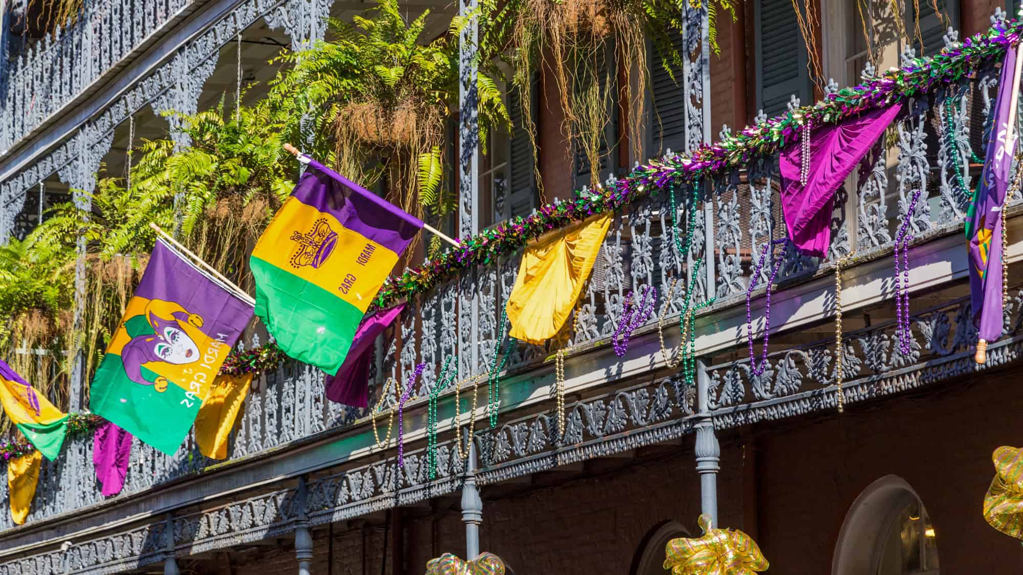 Colorful Mardi Gras flags, garlands, and beads decorate an ornate wrought-iron balcony with lush ferns above.