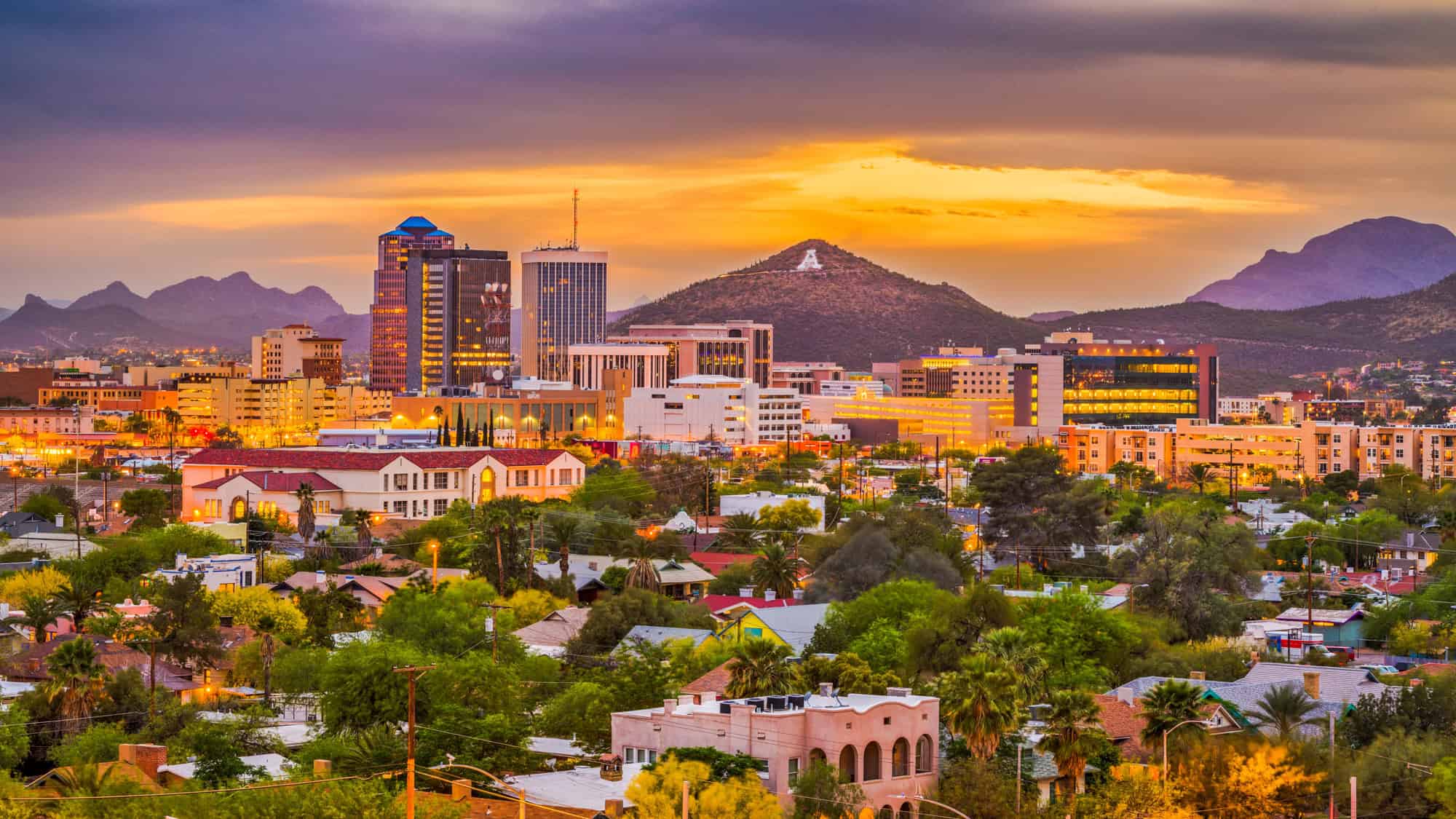 The Tucson city skyline glows under a golden sunset, with desert mountains in the distance.