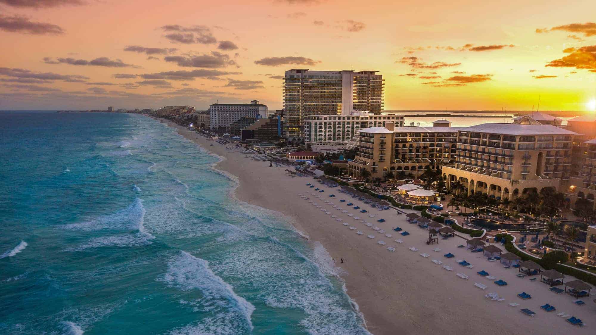 Aerial view of Cancun's beachside resorts at sunset. The ocean waves gently lap the sandy shore, while the skyline of hotels and buildings stretches along the coastline.