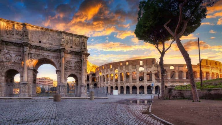 The image depicts the Arch of Constantine with the Colosseum in the background, bathed in warm sunset light under a dramatic sky with scattered clouds.