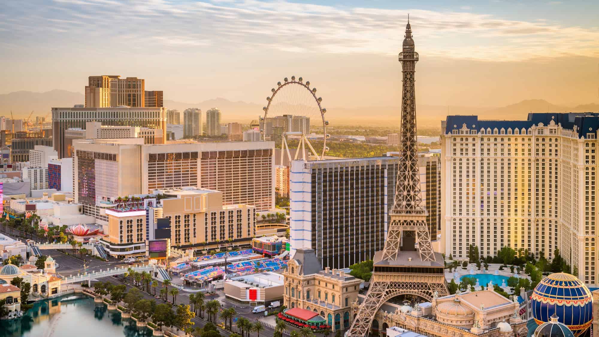 Daytime aerial view of the Las Vegas Strip, featuring the Eiffel Tower replica and the High Roller Ferris wheel, surrounded by hotels and casinos under a partly cloudy sky.