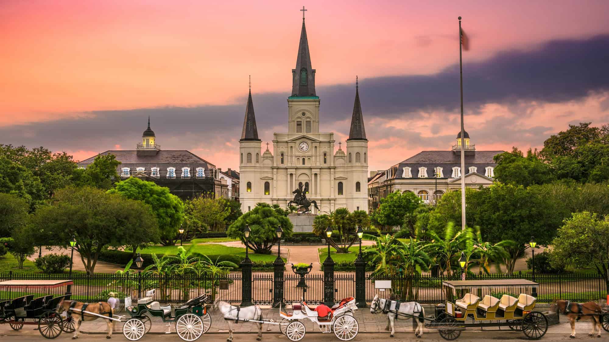 St. Louis Cathedral in New Orleans at dusk, with horse-drawn carriages in front and a colorful sunset sky in the background