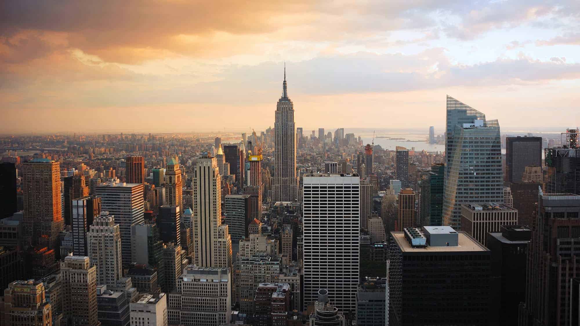 Aerial view of New York City at sunset, featuring the Empire State Building prominently in the center surrounded by various skyscrapers under a partially cloudy sky.