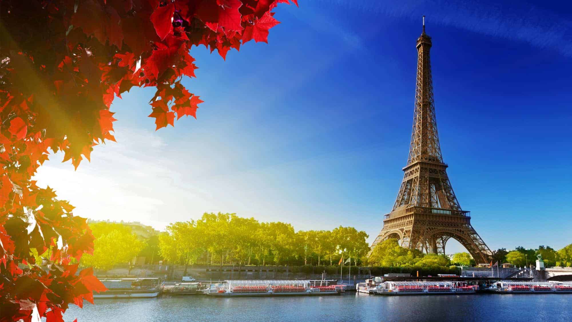 The Eiffel Tower seen from across the Seine River on a bright, sunny day. Red leaves frame the scene on the left, with tour boats docked along the river and green trees in the background.
