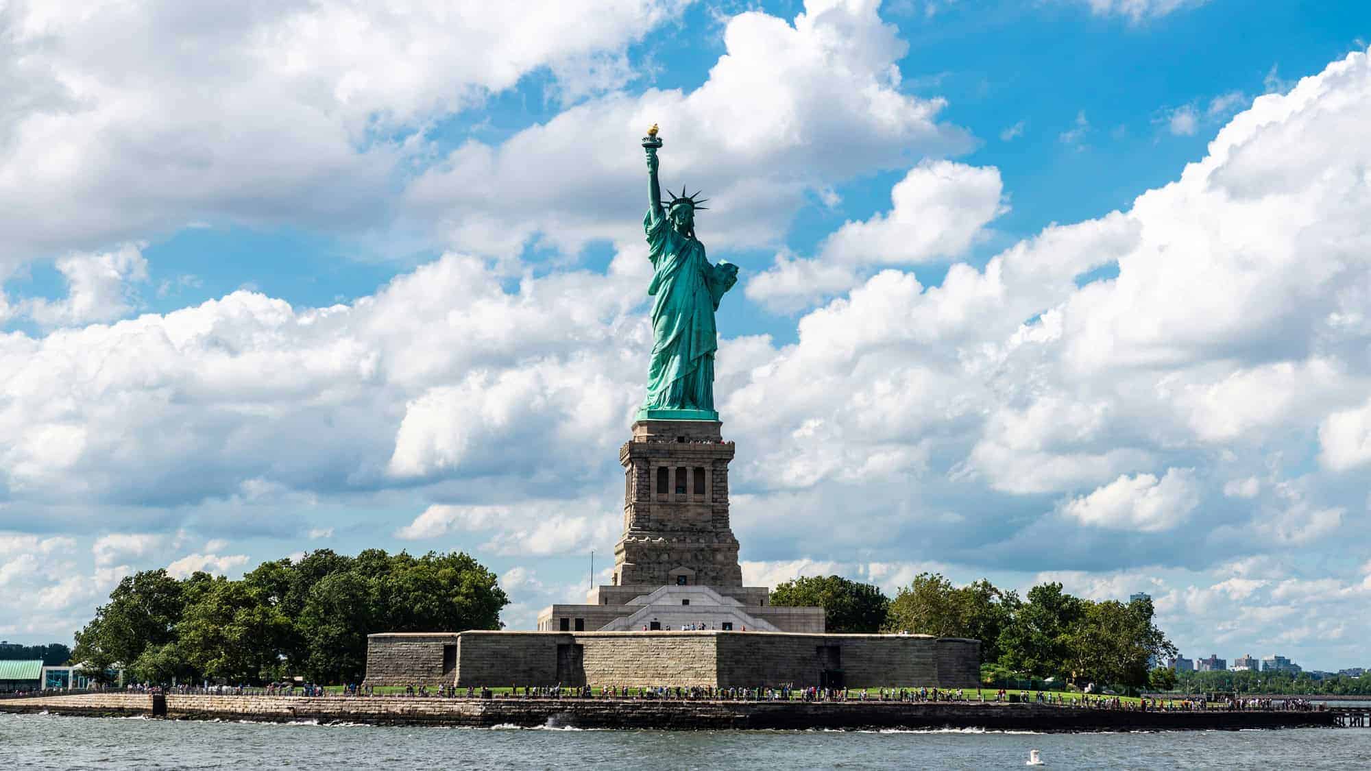 The image shows the Statue of Liberty on Liberty Island in New York Harbor, set against a backdrop of blue sky with fluffy white clouds.