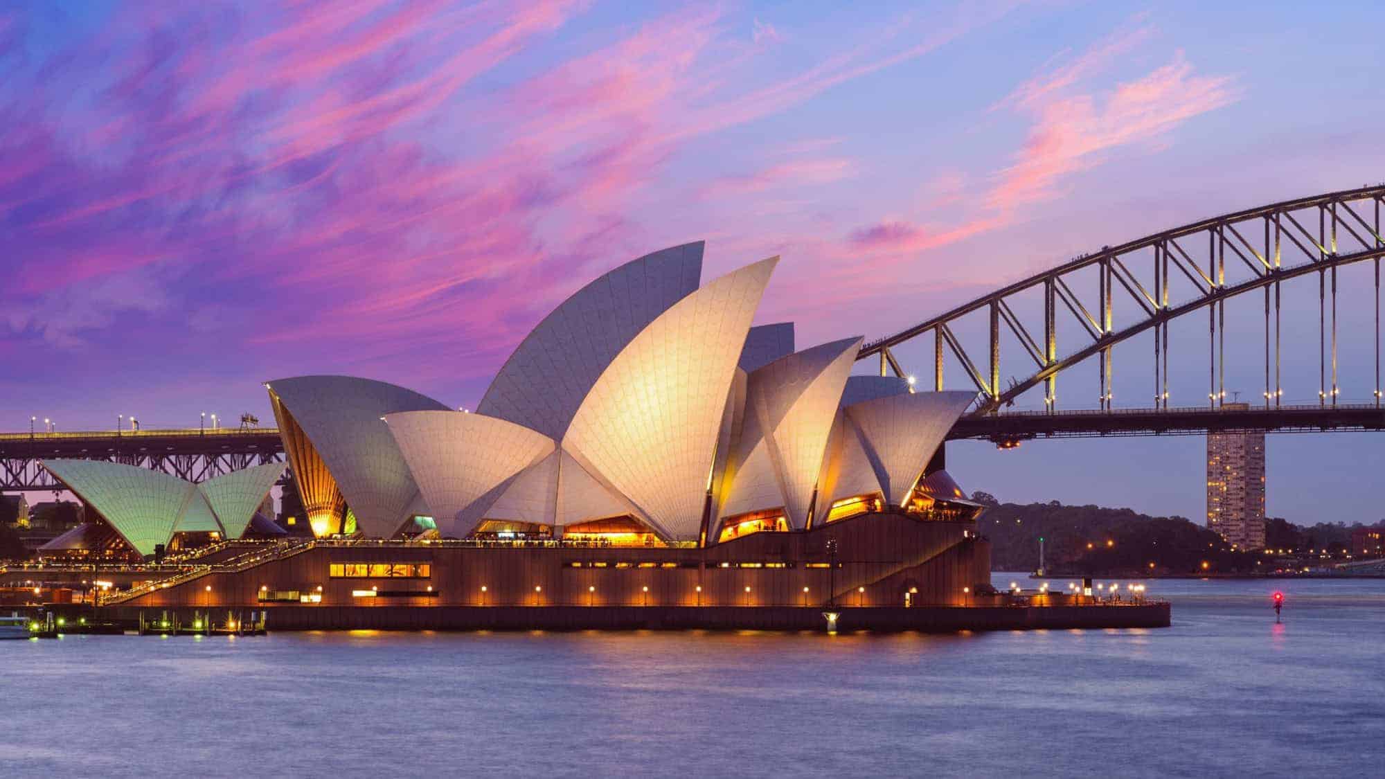 Sydney Opera House and Harbour Bridge at twilight with a vibrant purple-pink sky. The landmarks are illuminated, casting a warm glow over the harbor.