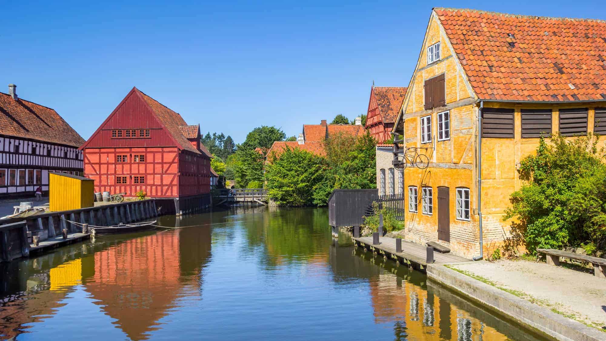Charming, colorful half-timbered houses with red and orange roofs reflect in the calm canal waters on a sunny day in Aarhus.