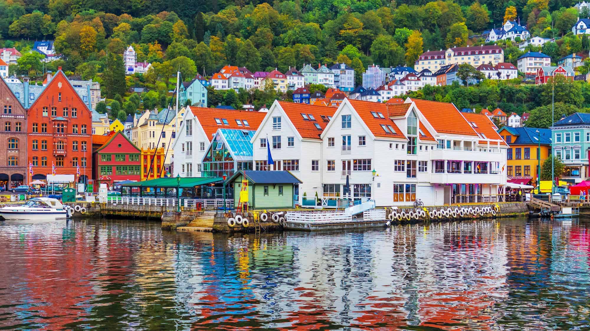 Colorful wooden houses line the waterfront in Bergen, reflecting vibrantly in the still water, with lush green hills rising behind them.