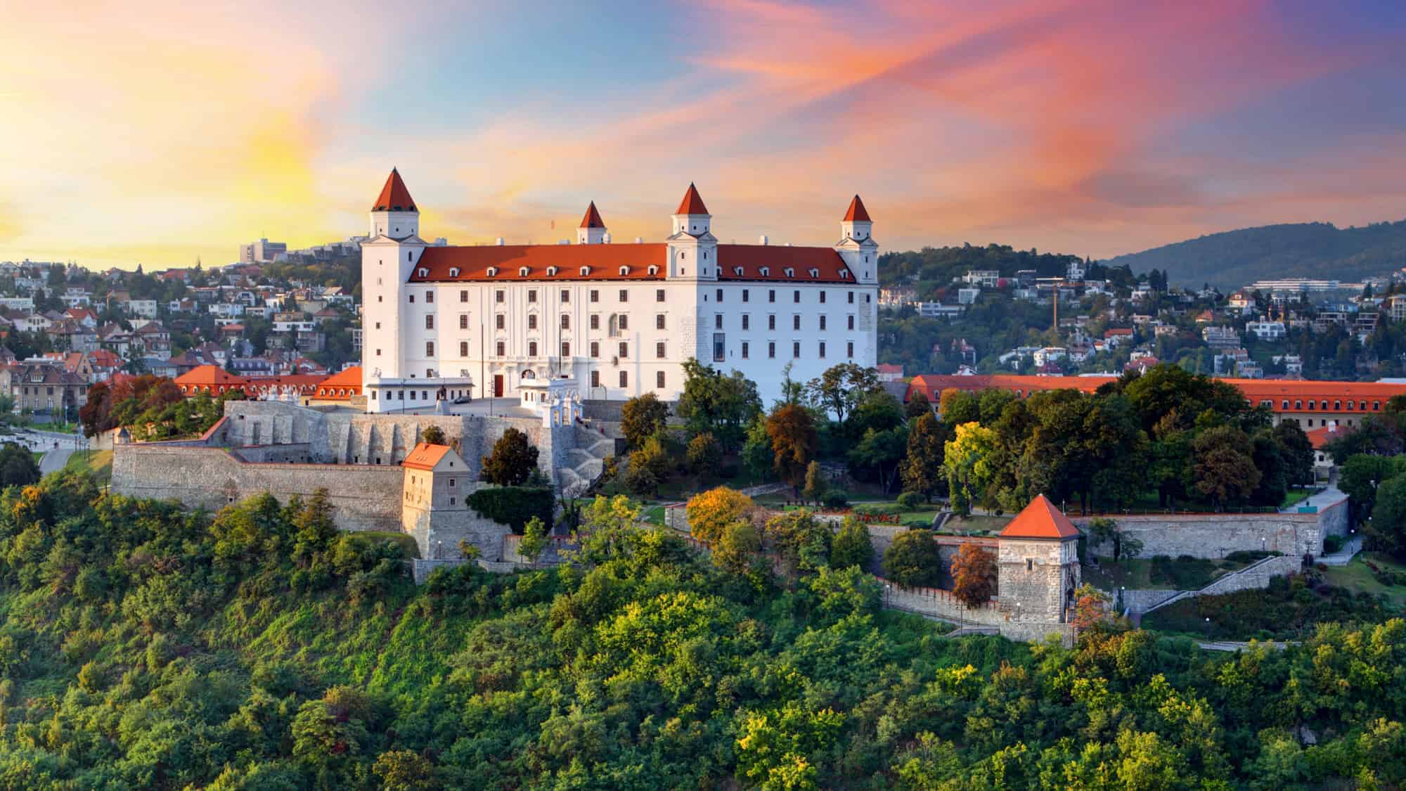 The majestic Bratislava Castle sits on a hill overlooking the city, bathed in the soft hues of a sunset sky.
