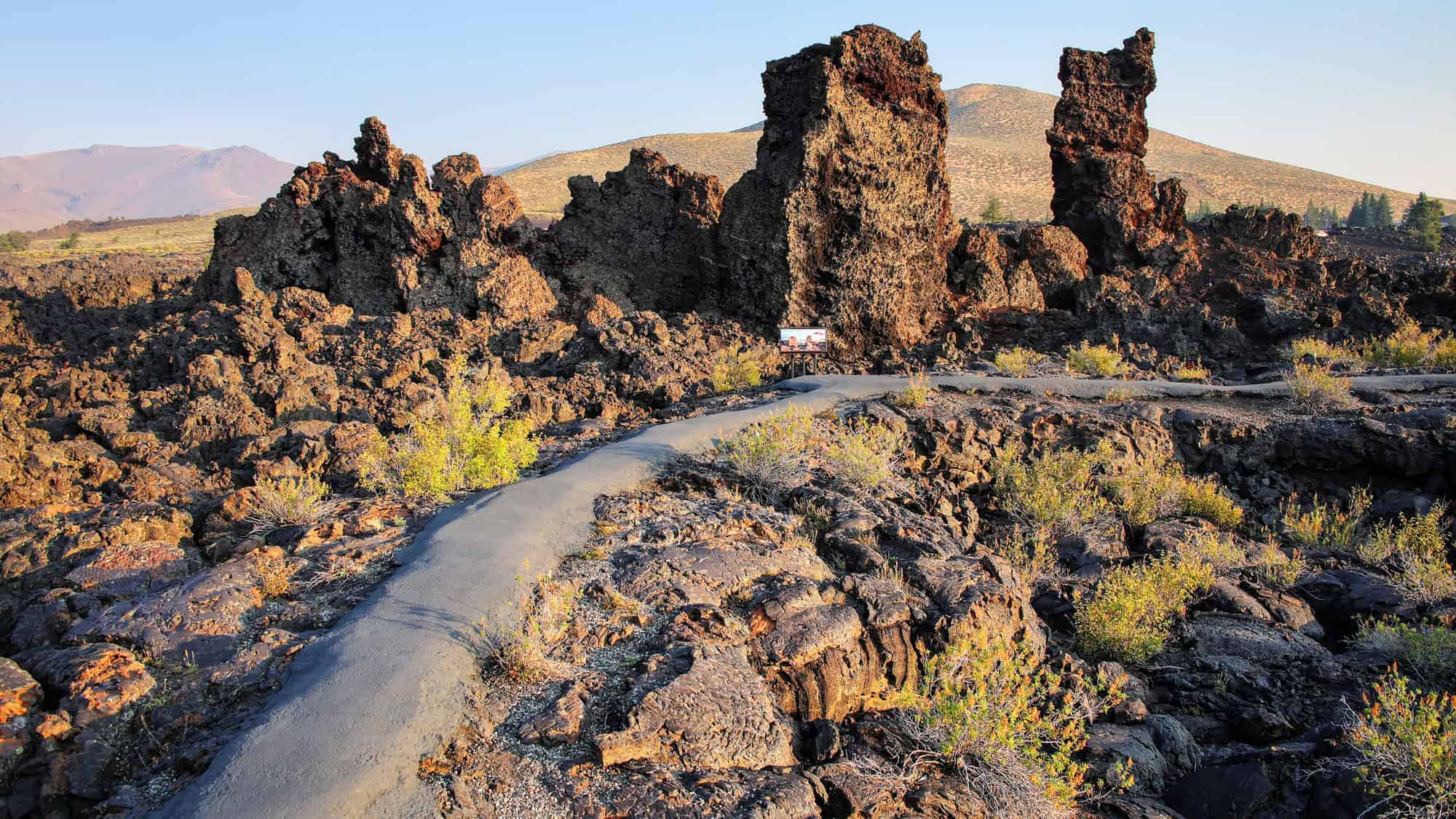 A paved path winds through rugged, dark volcanic rock formations at Craters of the Moon National Monument, Idaho.
