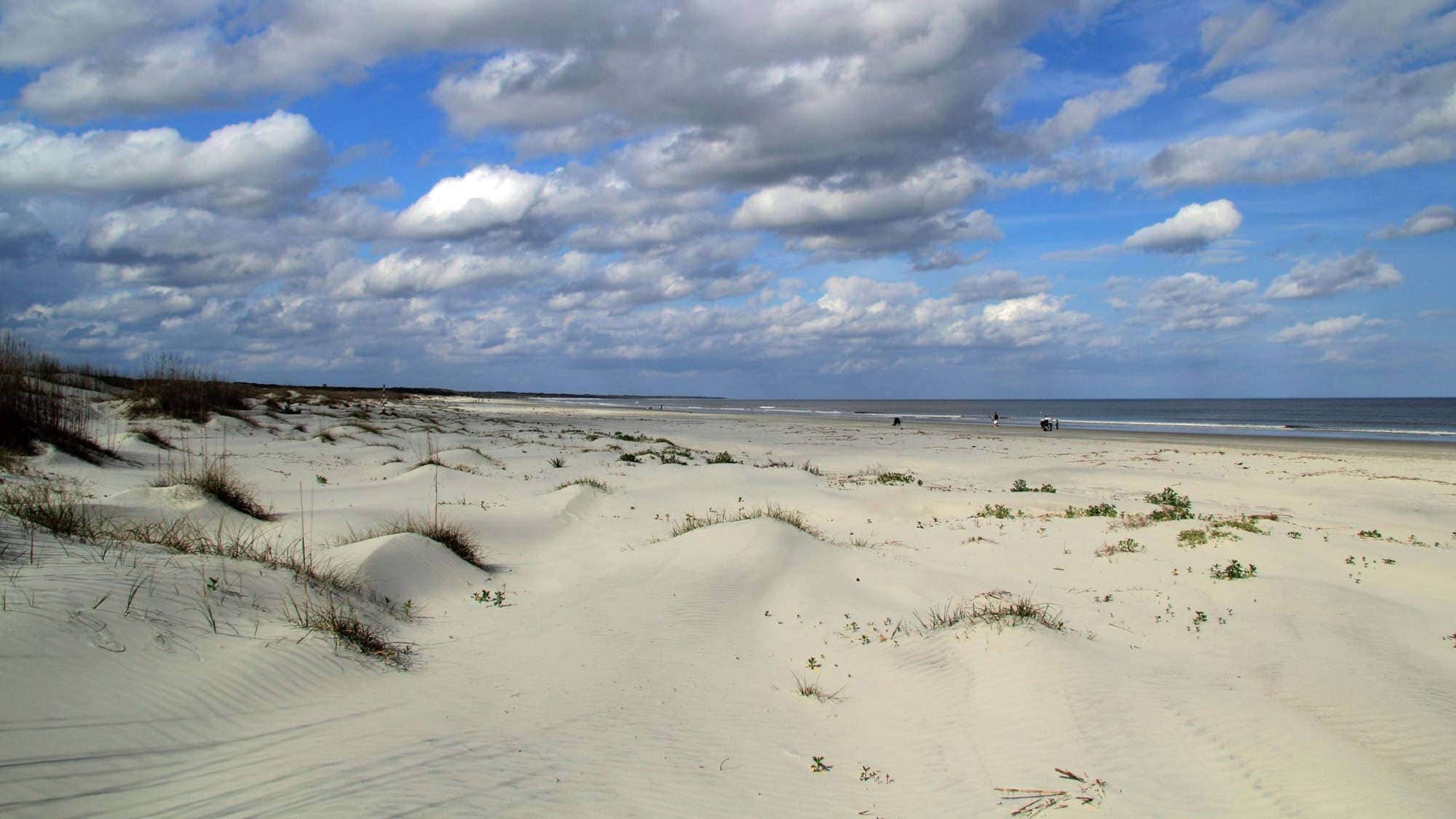 A vast, sandy beach with gentle dunes and sparse vegetation, under a partly cloudy sky, creating a serene and expansive seascape.