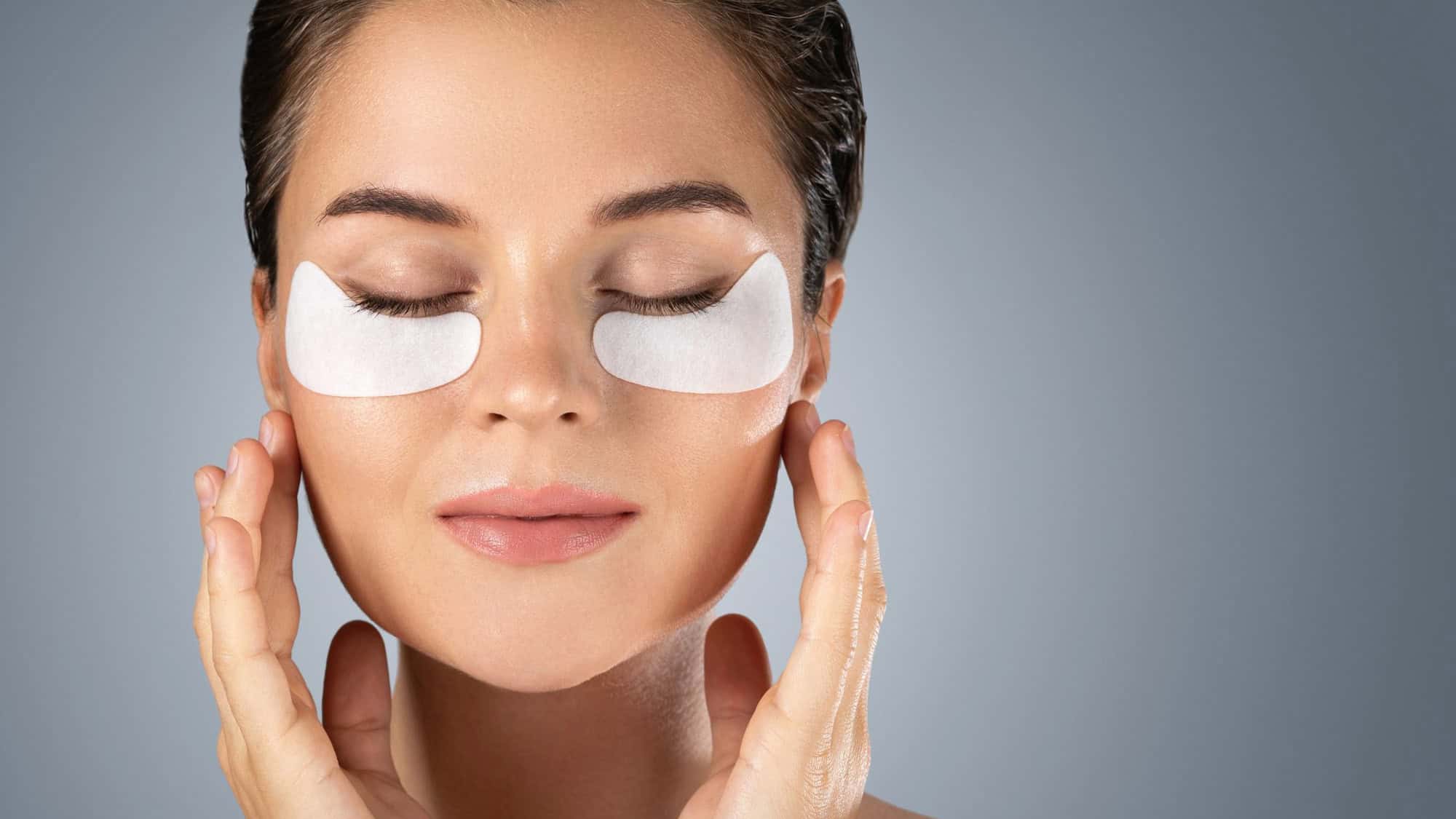 Woman with under-eye patches and closed eyes, gently touching her face.
