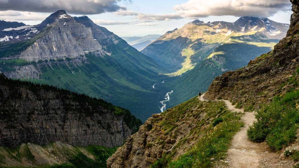 A narrow trail winds along a steep cliffside in Glacier National Park, offering a breathtaking view of lush, green valleys and towering mountain ranges.
