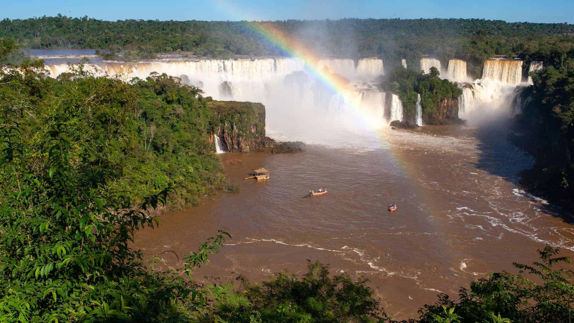 A breathtaking scene of Iguazu National Park with powerful waterfalls plunging into a river, framed by lush rainforest and highlighted by a vivid rainbow arcing across the sky.