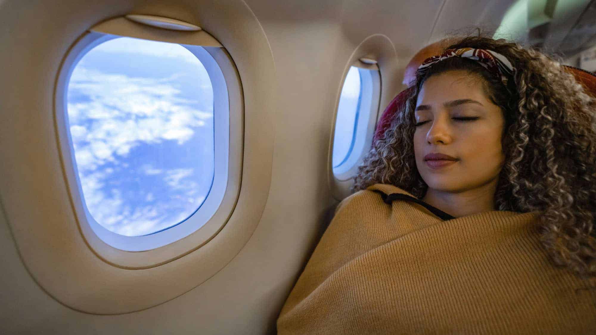 A woman with curly hair, wrapped in a blanket, is peacefully sleeping in an airplane seat next to a window showing a cloudy sky.