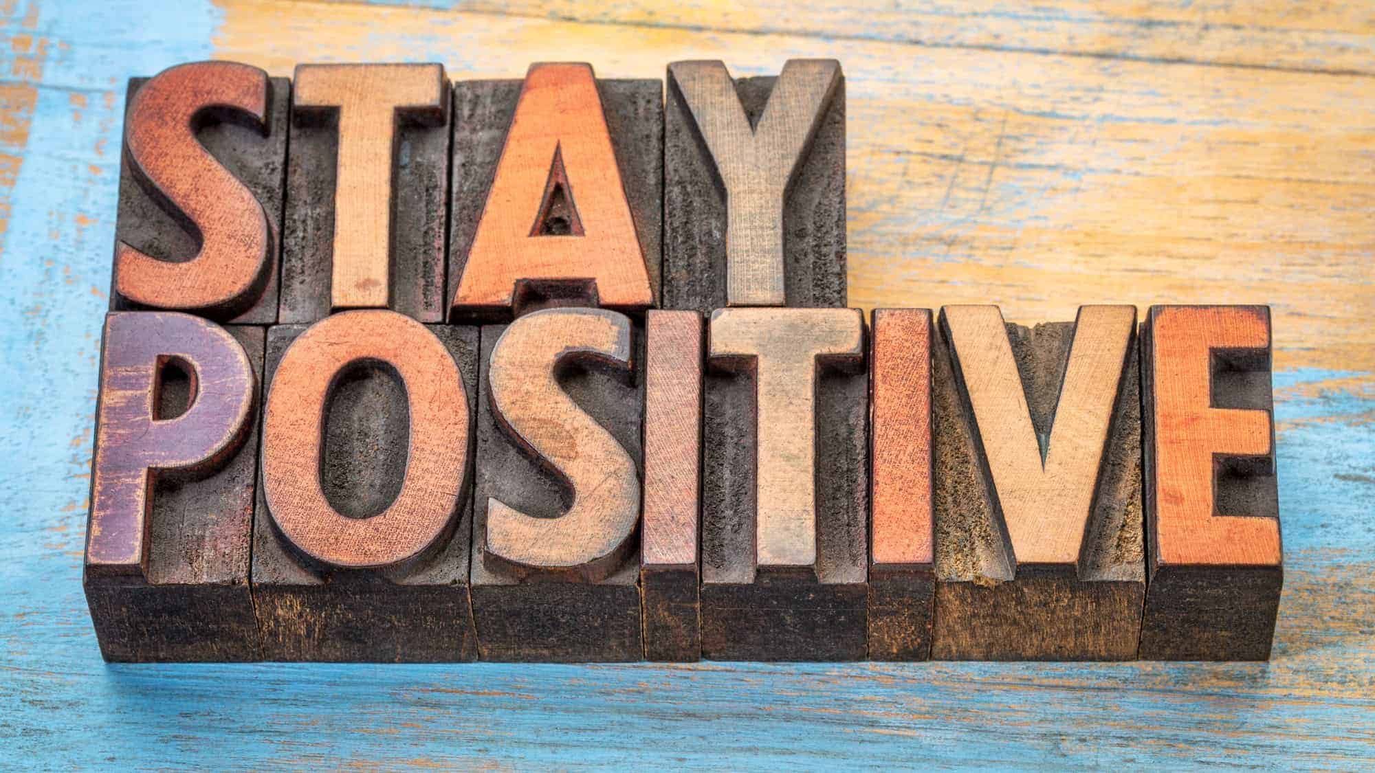 A colorful wooden block sign that reads "STAY POSITIVE" with varying shades and textures on a painted wooden surface.