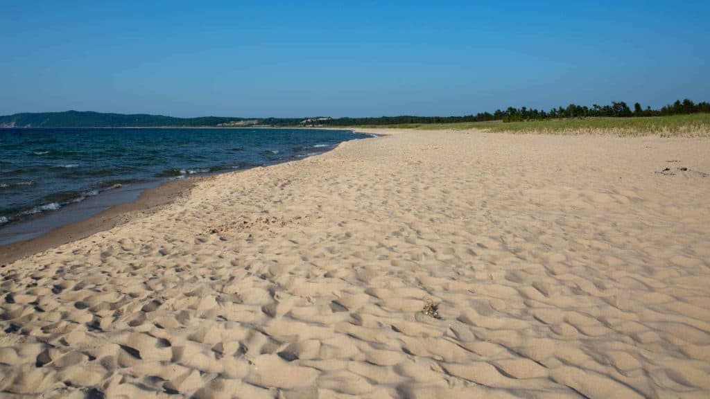 A sandy beach with gentle waves and an expansive view of Lake Michigan, bordered by grassy dunes under a clear blue sky.