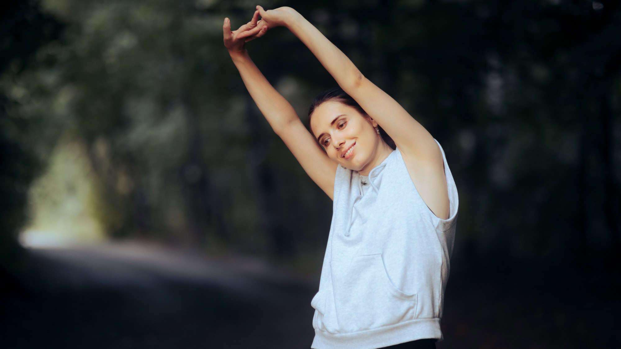 A woman in a light gray sleeveless hoodie stretching her arms above her head while standing on a forest path, looking content.
