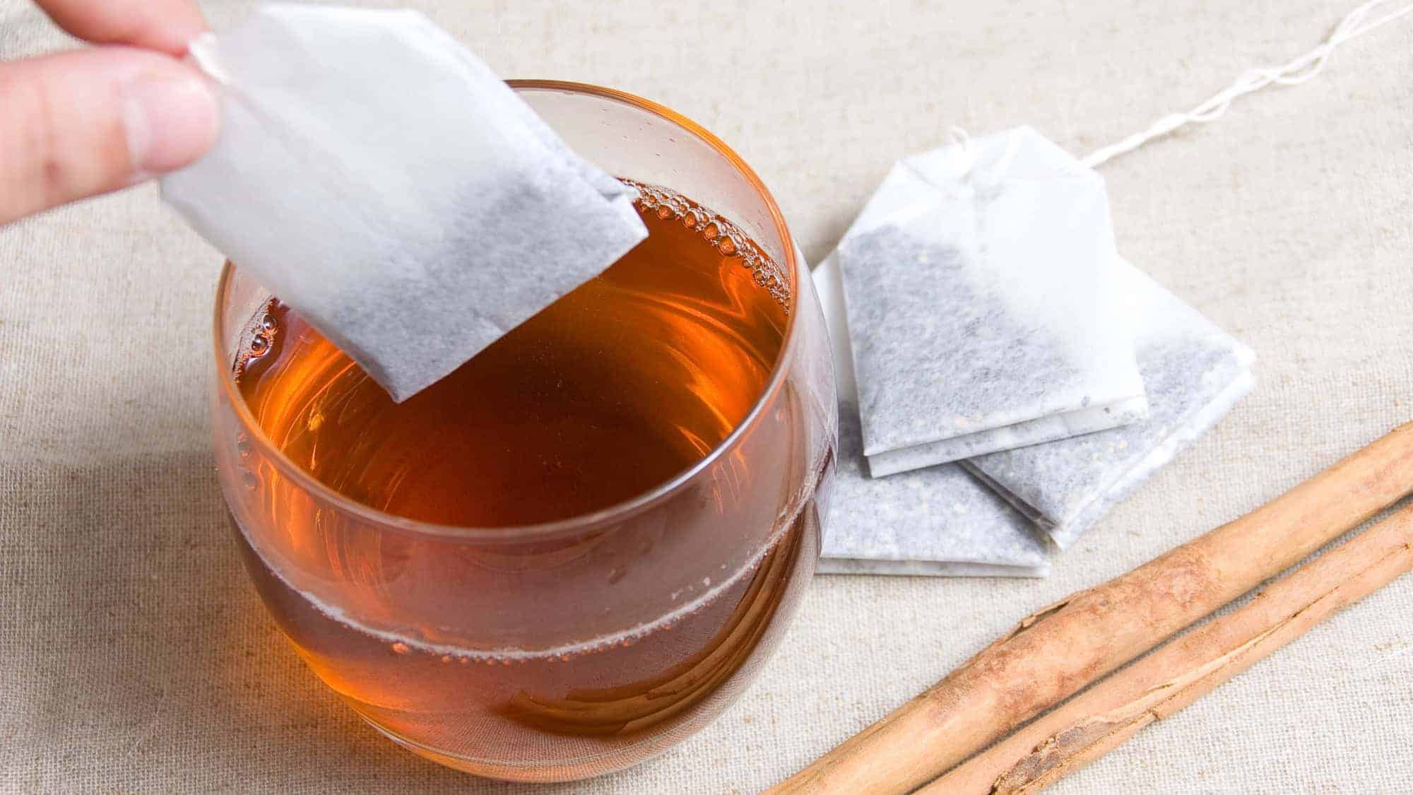 A hand holding a tea bag above a glass cup filled with tea, with additional tea bags and a cinnamon stick on the table.