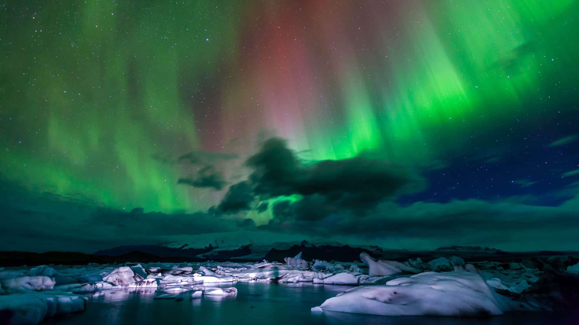 The vibrant Northern Lights dance in the night sky above icy waters and snow-covered landscape in Vatnajökull National Park, Iceland.