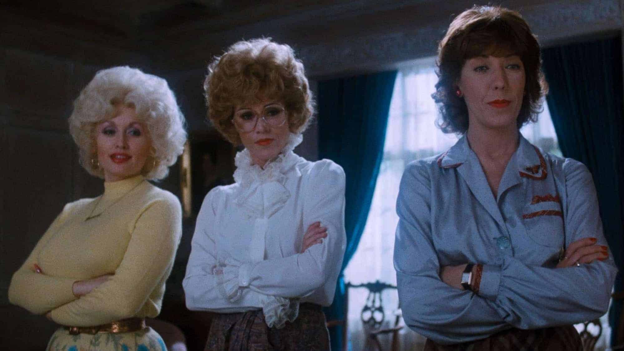 Three women with determined expressions stand together, arms crossed. They are dressed in 1980s office attire, and their stances convey confidence and solidarity.
