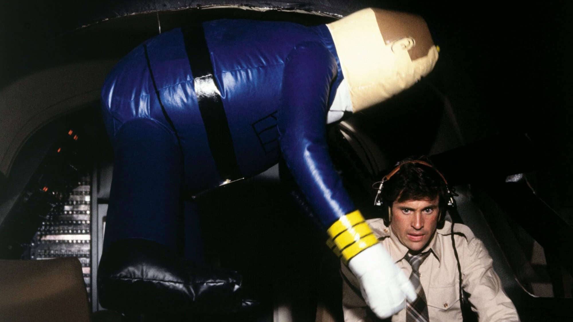 A comedic scene with a pilot seated in the cockpit next to a large, inflatable co-pilot figure. The pilot looks serious while the inflatable figure is positioned humorously.