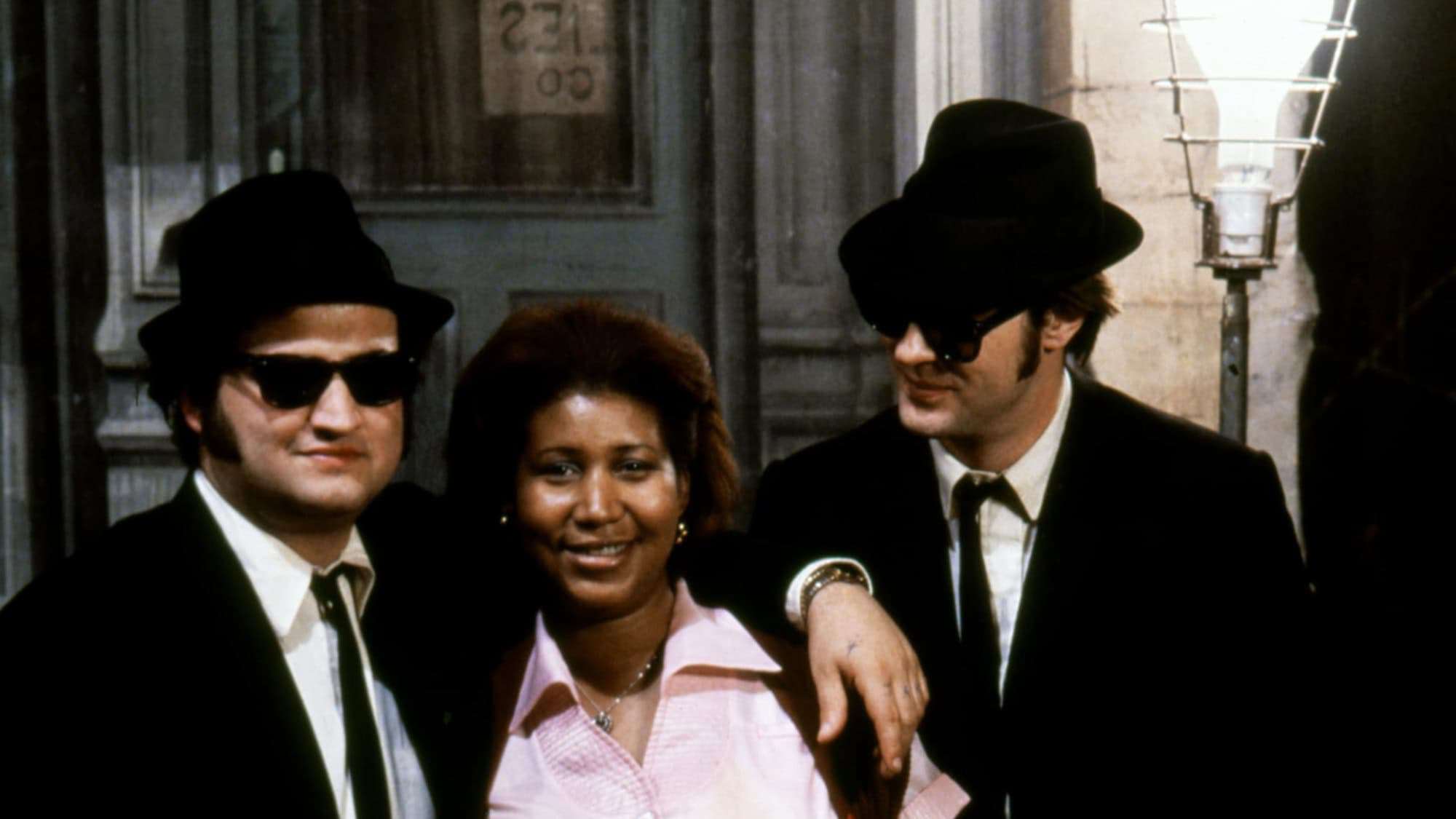 Two men dressed in black suits, hats, and sunglasses stand next to a woman. They are smiling and have a relaxed demeanor, with one man’s arm around the woman.