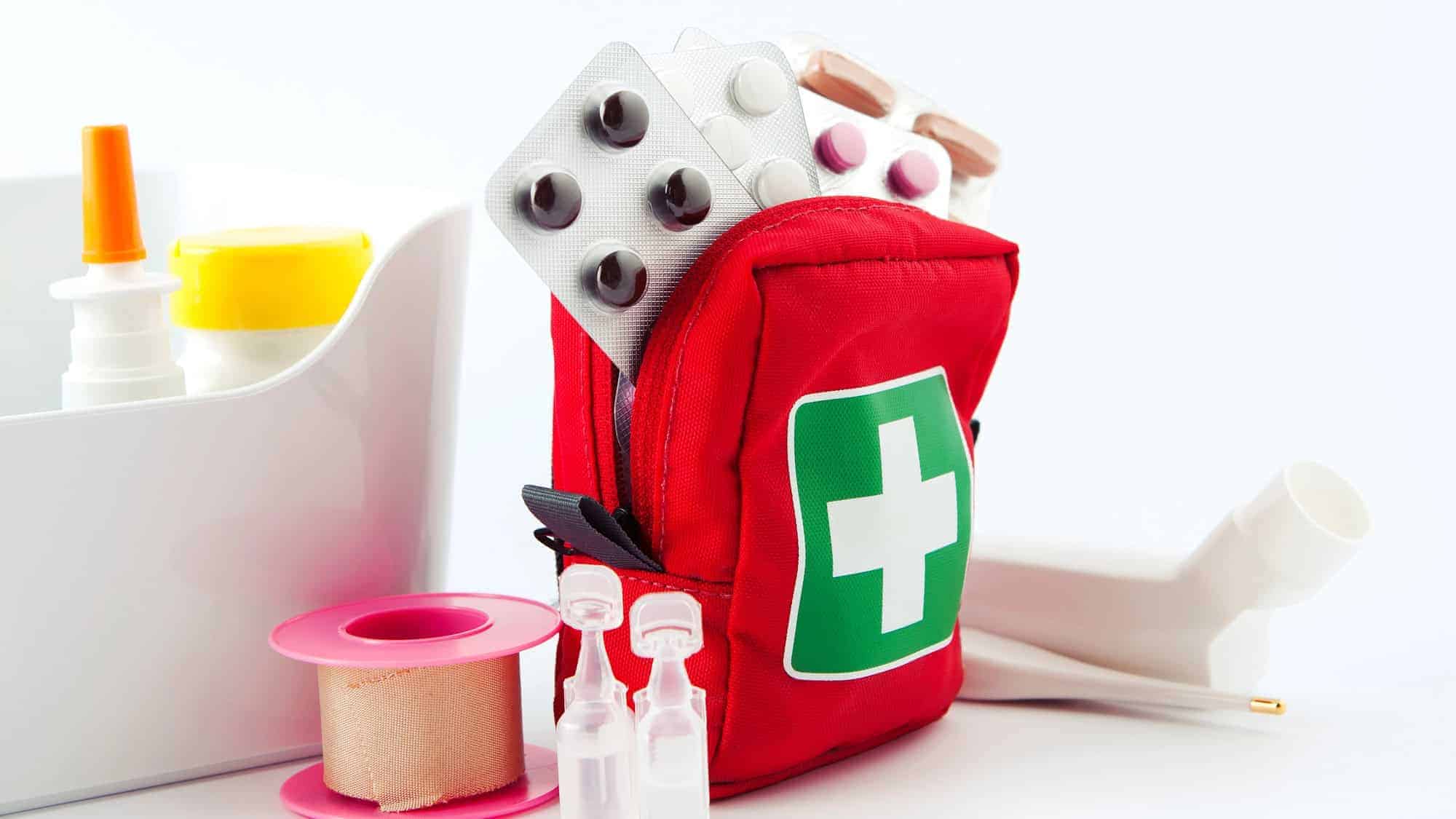 A red first aid kit with a green cross symbol, filled with various medical supplies like blister packs of pills, a thermometer, and nasal spray, surrounded by bandages and other first aid items.