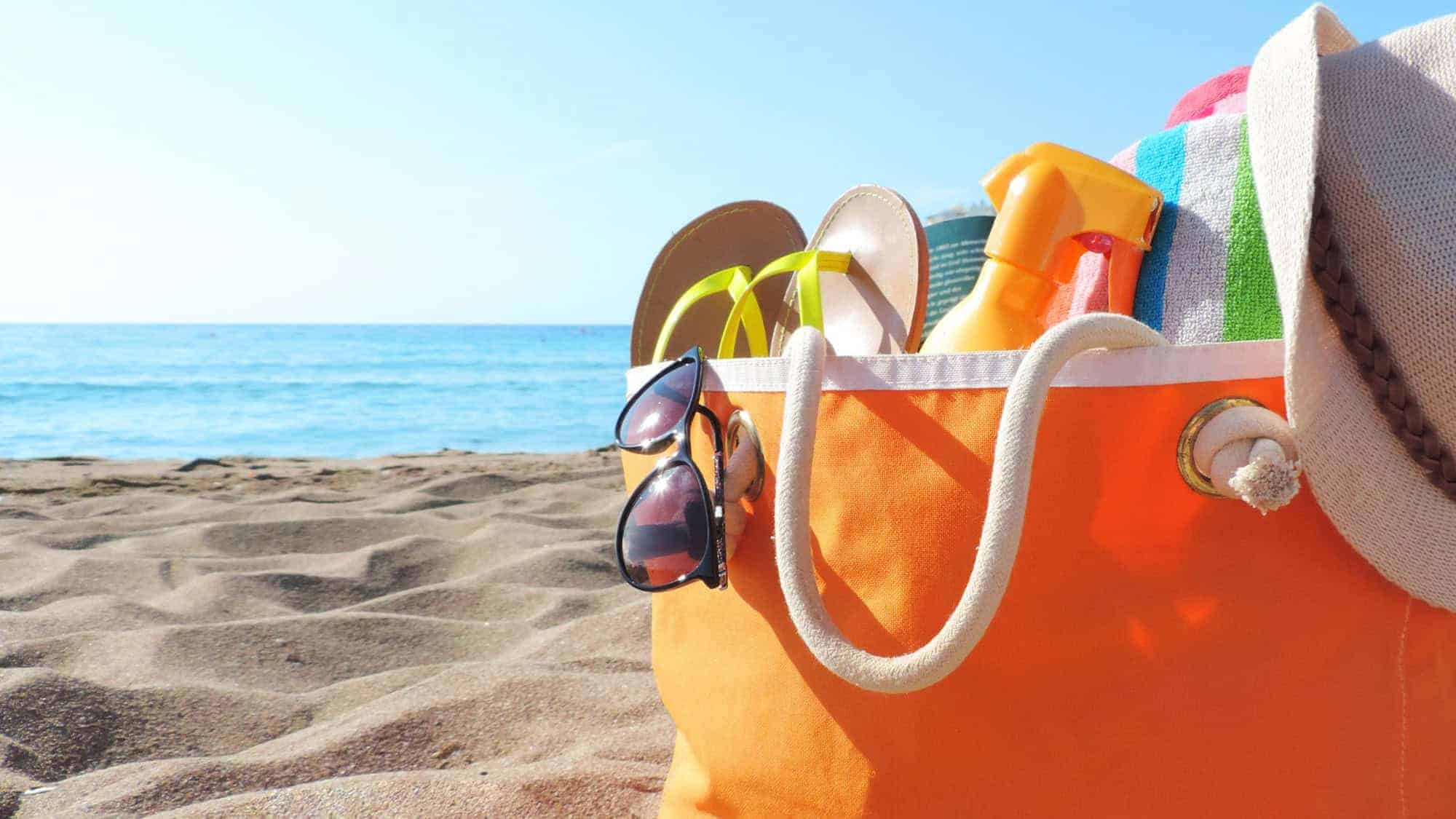 An orange beach bag sitting on the sand, filled with summer essentials like flip-flops, sunglasses, sunscreen, and a towel, with the ocean in the background.