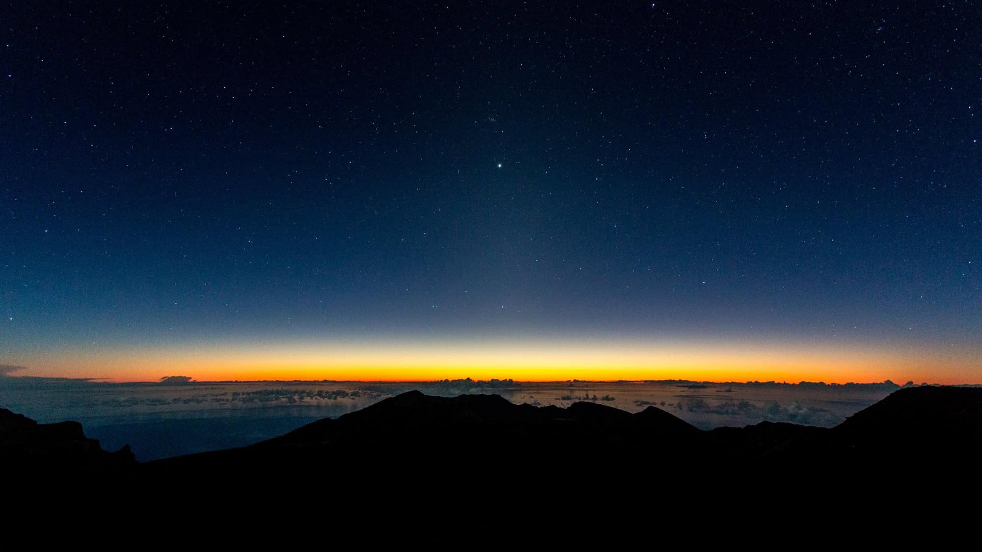 A breathtaking view from Haleakalā, with a clear night sky filled with stars and the first light of dawn beginning to illuminate the horizon.