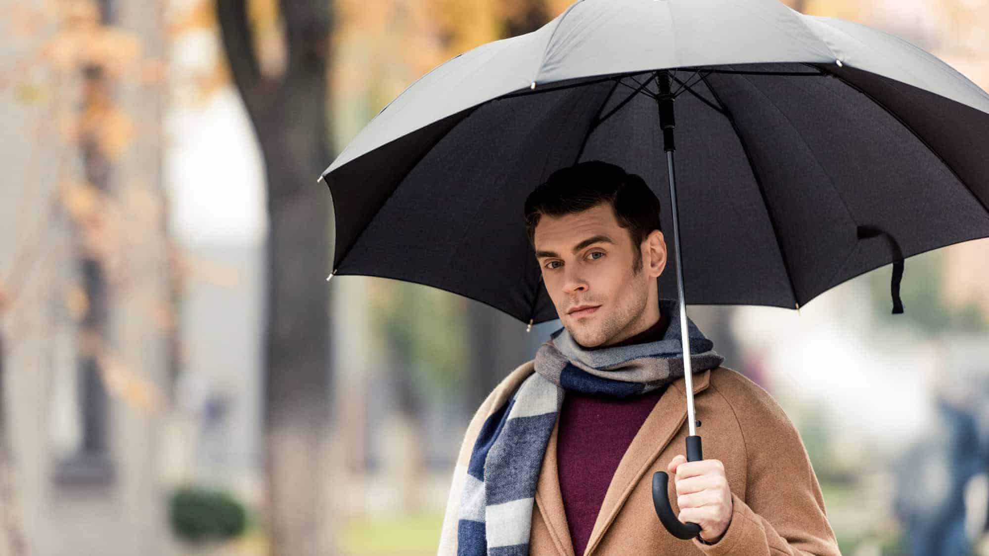 A man wearing a brown coat and scarf, holding a black umbrella while standing outdoors in a fall setting.