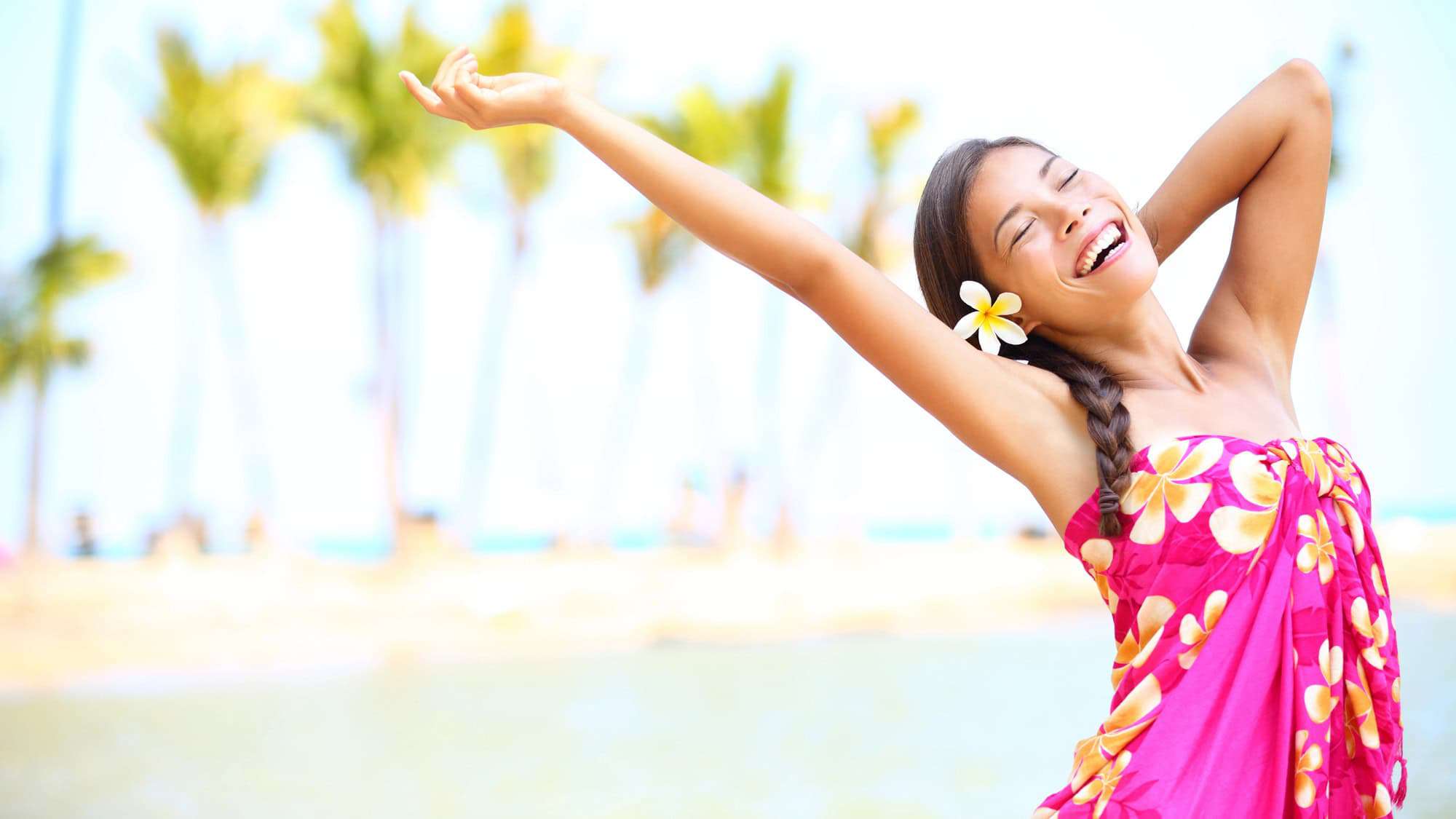 A happy woman in a vibrant pink dress with a flower in her hair, stretching her arms joyfully on a sunny beach with palm trees in the background.