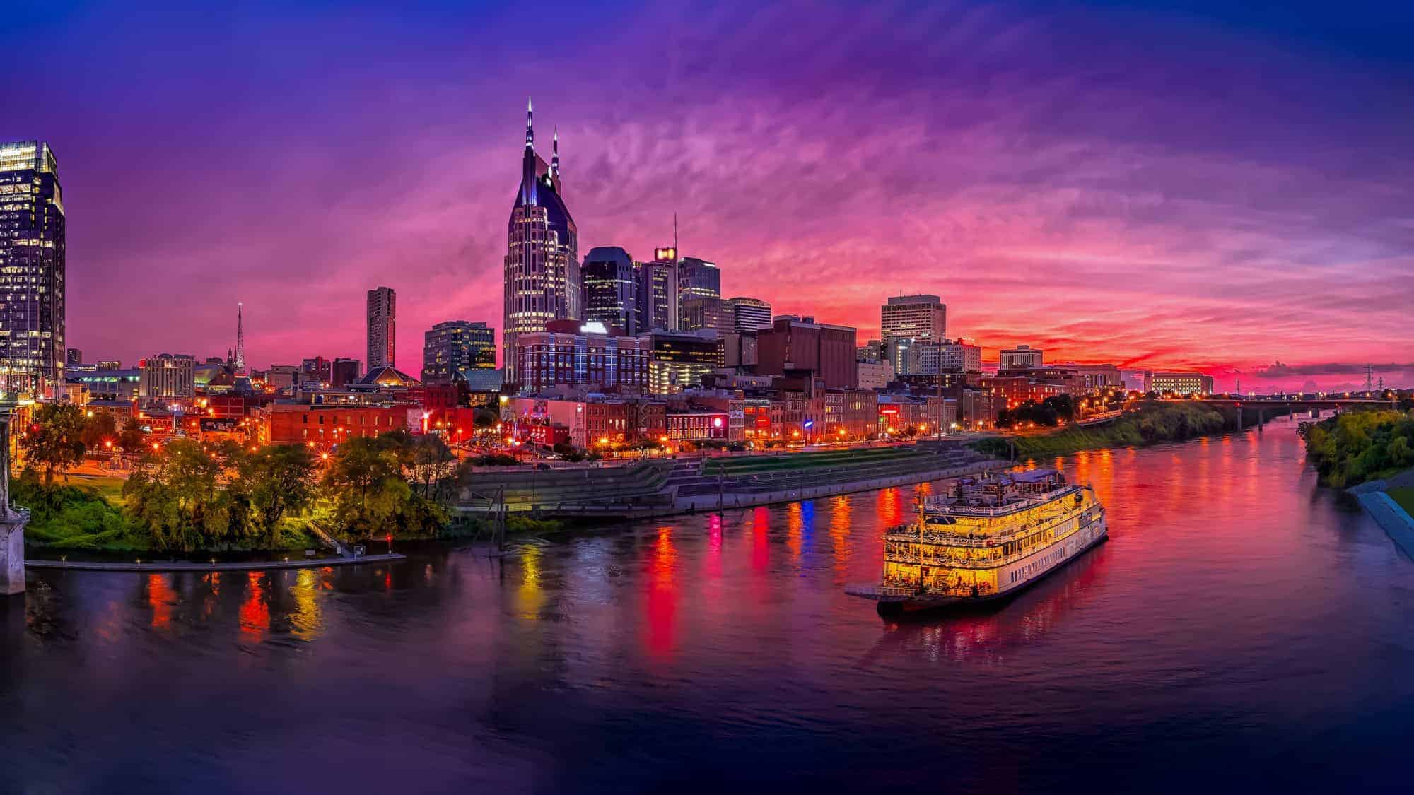 A vibrant panoramic view of Nashville's cityscape during sunset, with the skyline illuminated by colorful lights reflecting off the river. A riverboat is also visible on the water, adding a touch of charm to the bustling city scene.