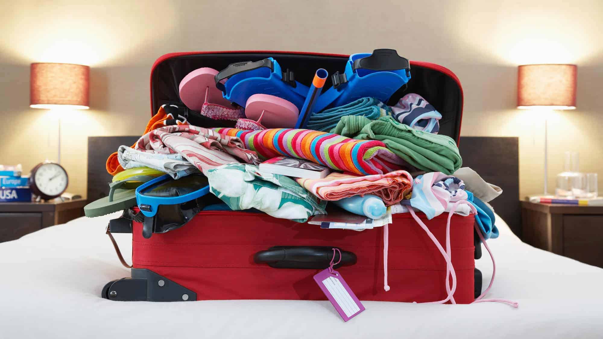 A red suitcase overstuffed with various colorful clothes and travel items, sitting on a white hotel bed with lamps and a clock in the background.