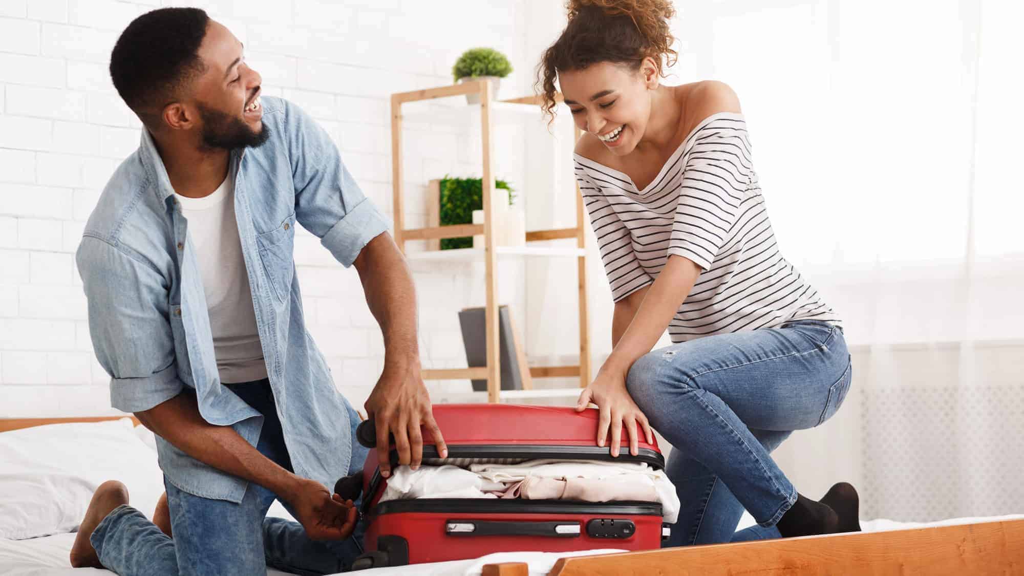A smiling couple trying to close an overfilled red suitcase on a bed, with both of them pressing down on the luggage.