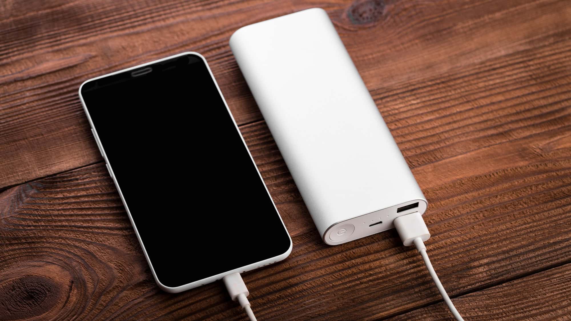 A smartphone connected to a white portable charger with a USB cable, resting on a wooden surface.