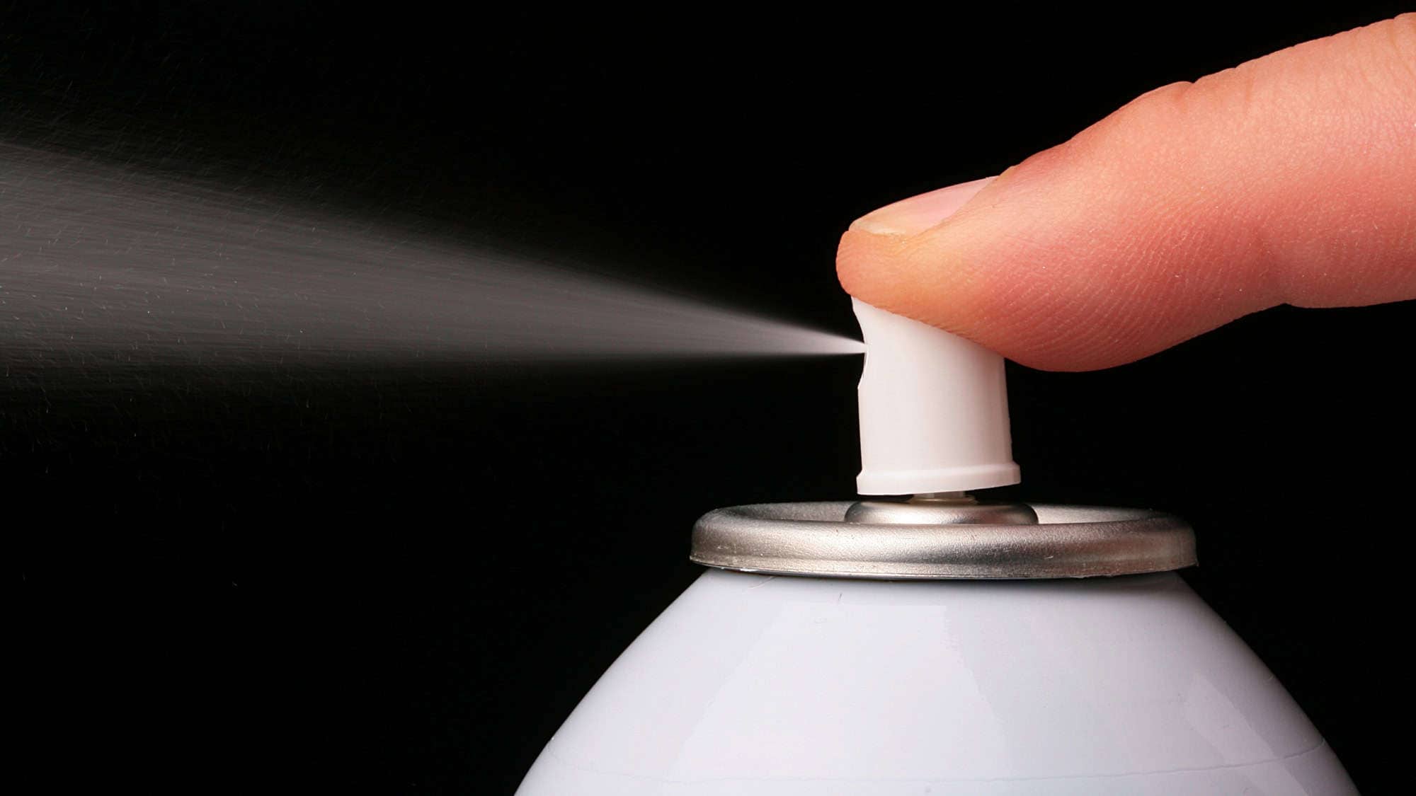 A finger pressing down on the nozzle of a spray can, releasing a mist of spray against a black background.