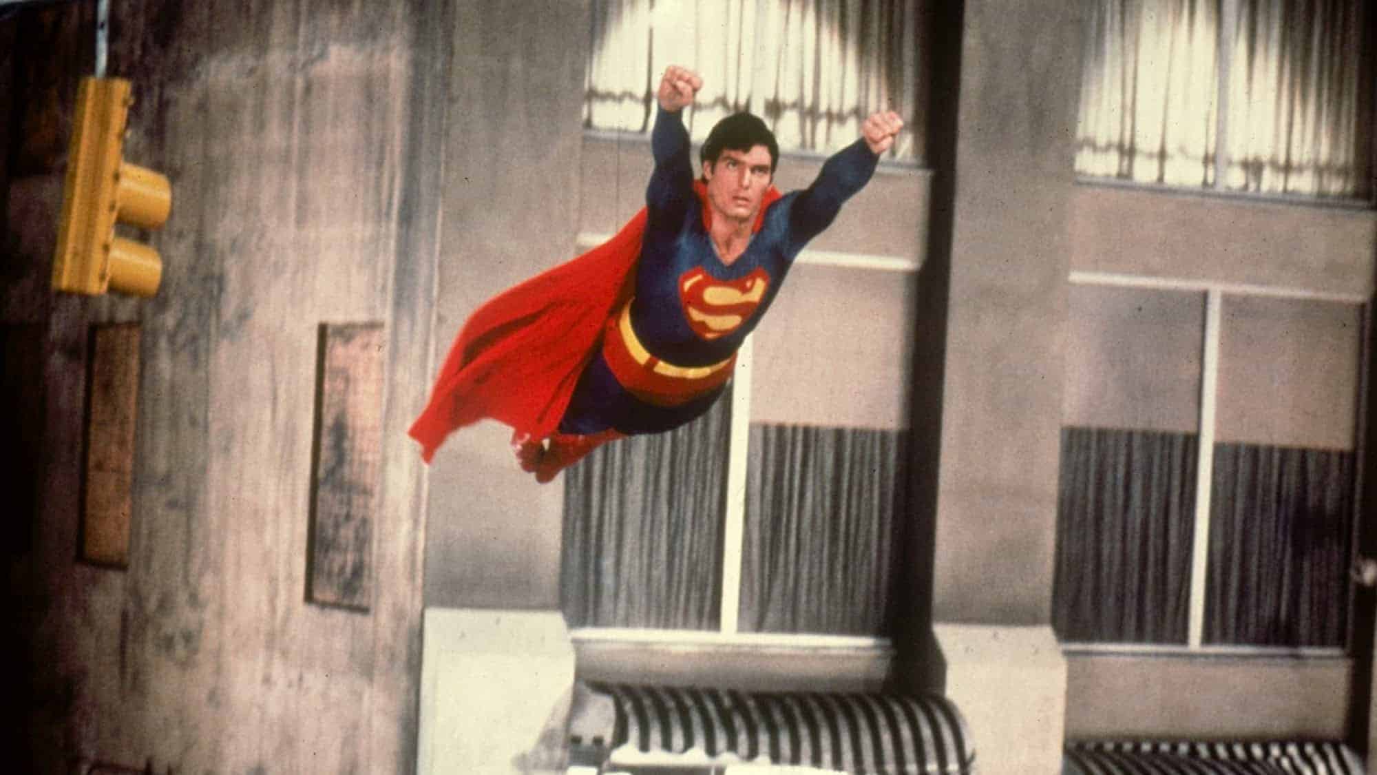 Superman is flying through the air in his iconic blue suit with a red cape, captured mid-flight above a cityscape.