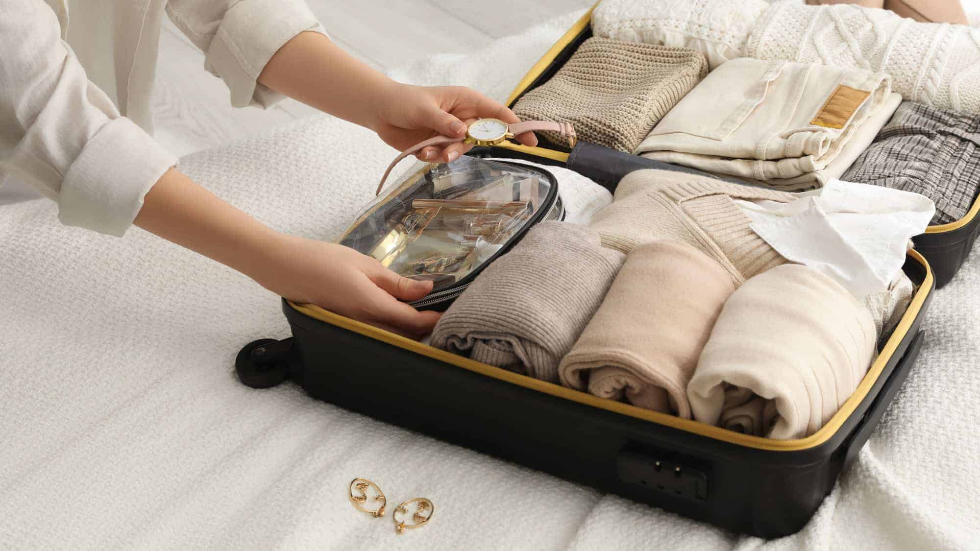 A person carefully placing a watch into a clear plastic pouch inside a neatly packed suitcase filled with folded clothing items, preparing for travel.