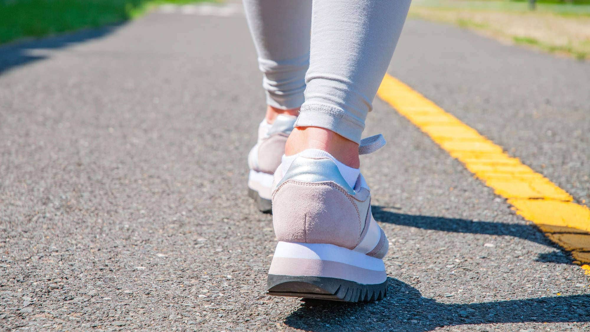 Close-up view of a person’s feet wearing sneakers, walking on a paved path with a yellow line.