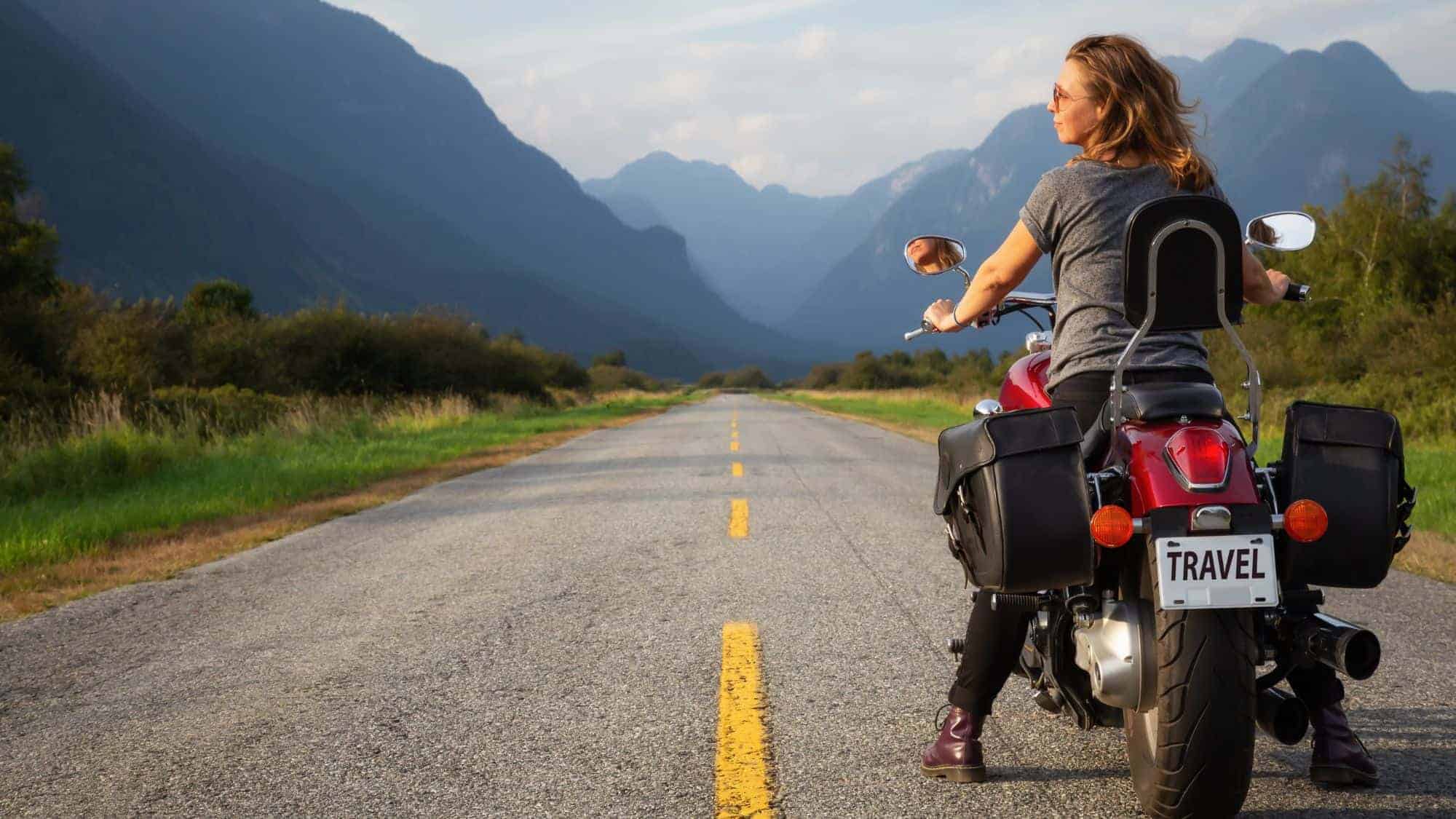 A woman sits on a red motorcycle parked on an open road. Majestic mountains and greenery surround the landscape in the distance.