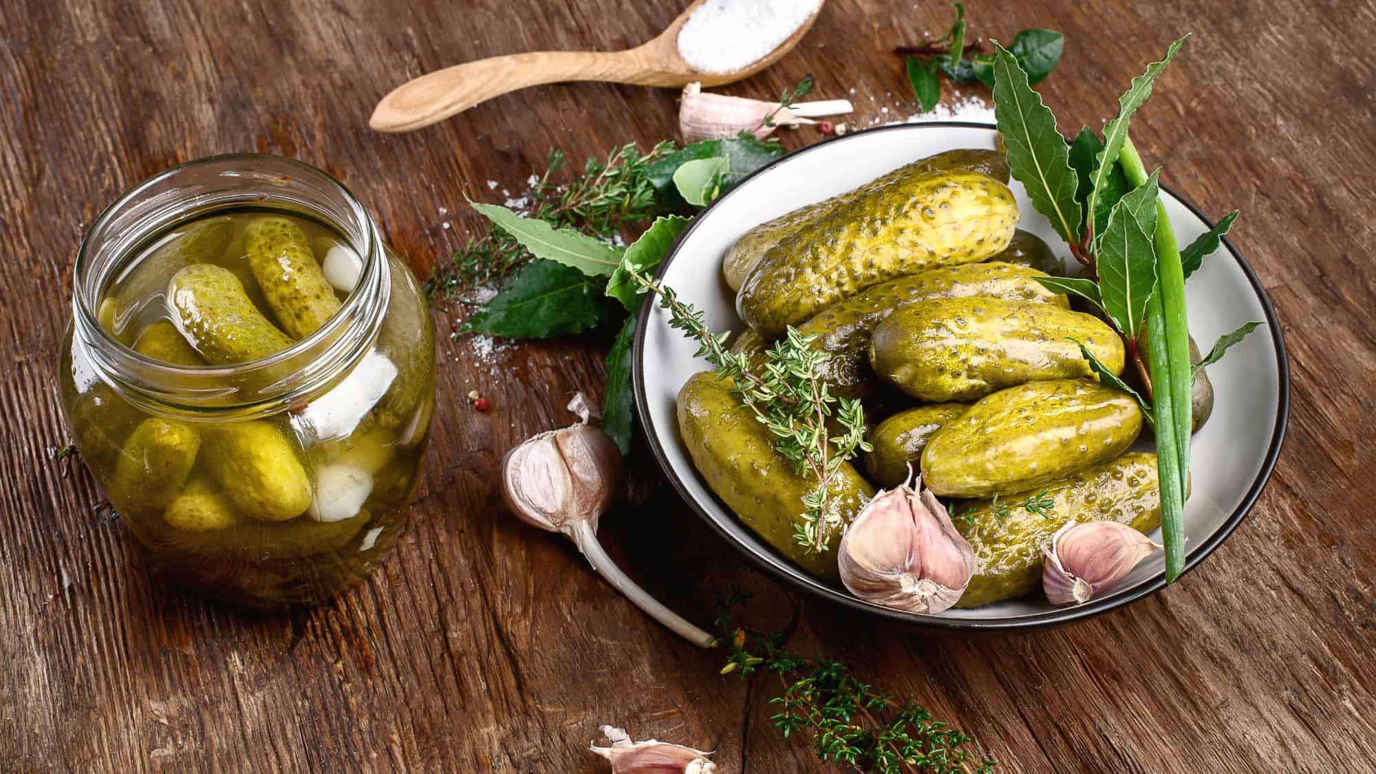 A bowl of pickles sits on a wooden table, accompanied by garlic cloves and fresh herbs. A jar of pickles is placed beside the bowl.