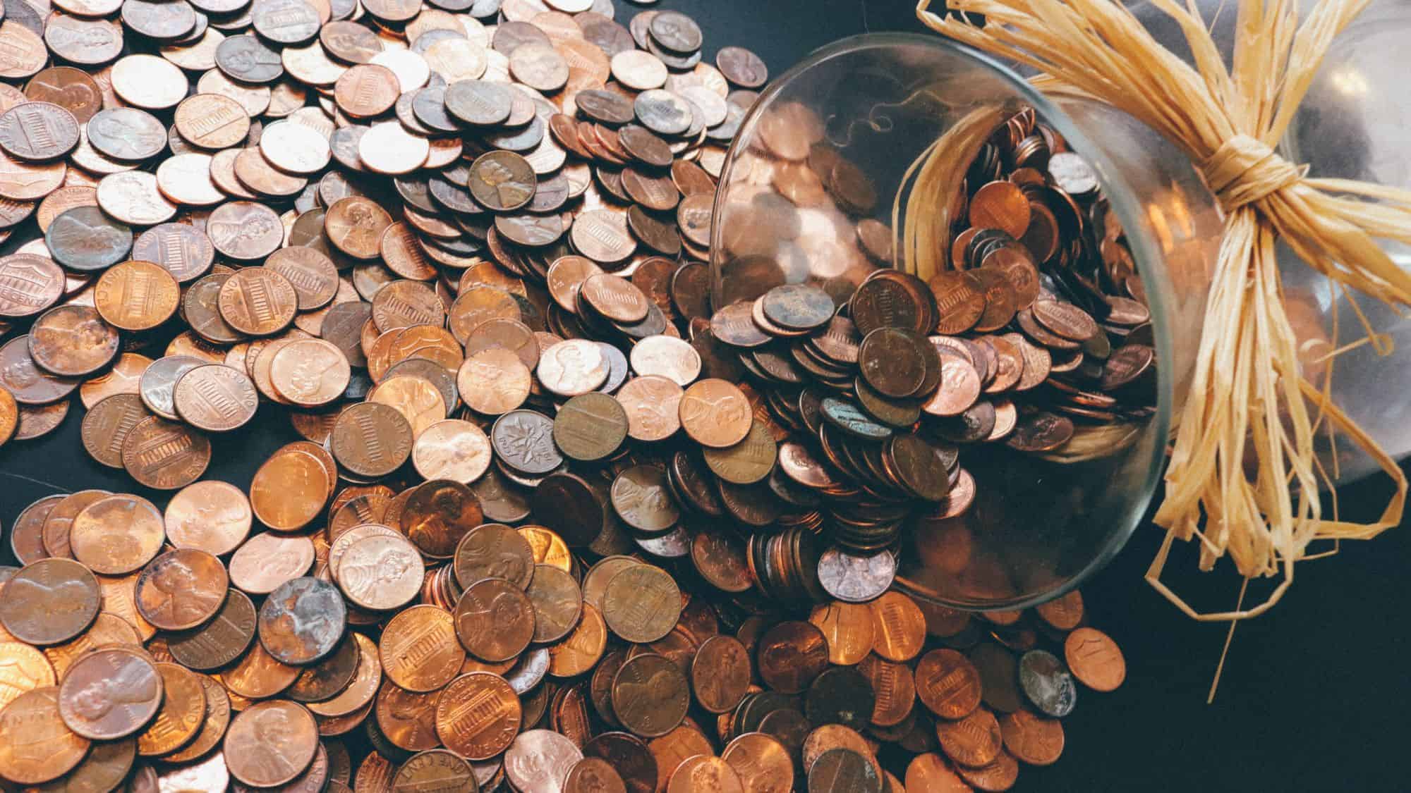 A glass jar tipped over, spilling a large pile of pennies across a dark surface, creating a cascade of copper coins.