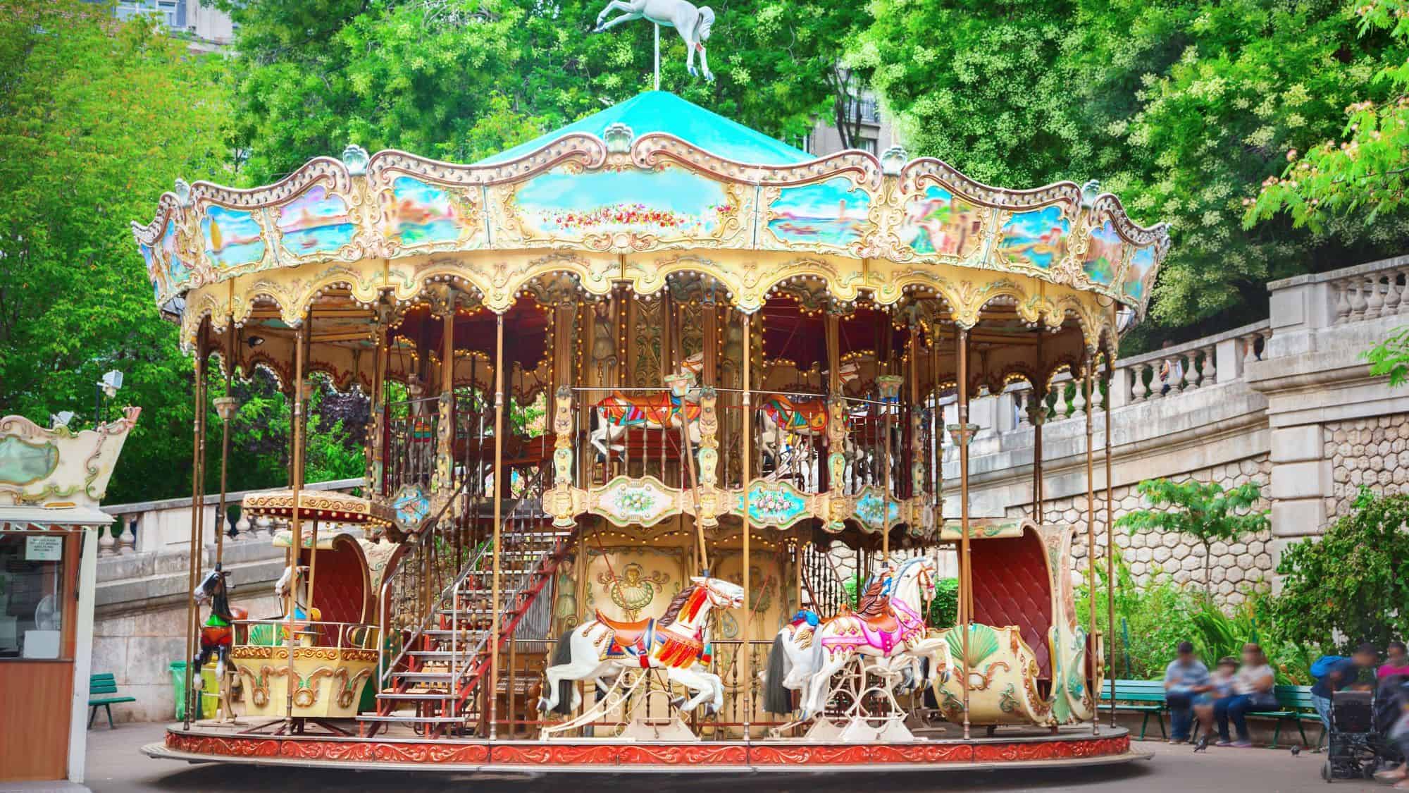 A colorful and ornate merry-go-round with intricately painted horses and lights, set in a park surrounded by green trees.